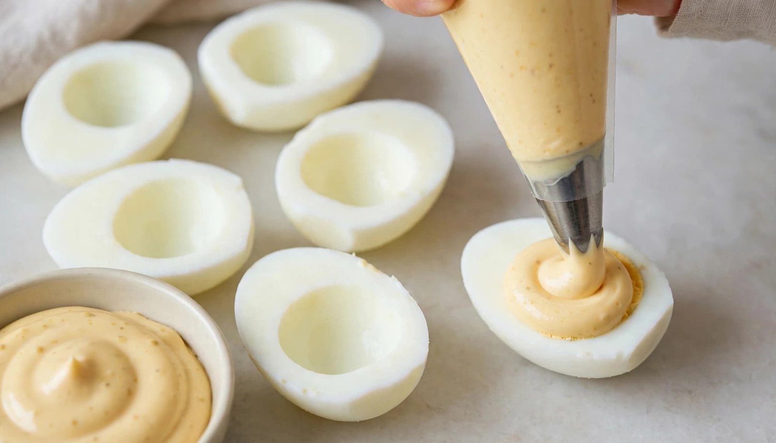 A piping bag fills an egg white beside five more whites and a bowl of creamy filling.