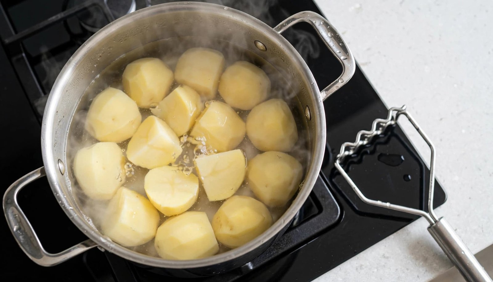 Peeled potatoes simmer in a saucepan beside a potato masher.