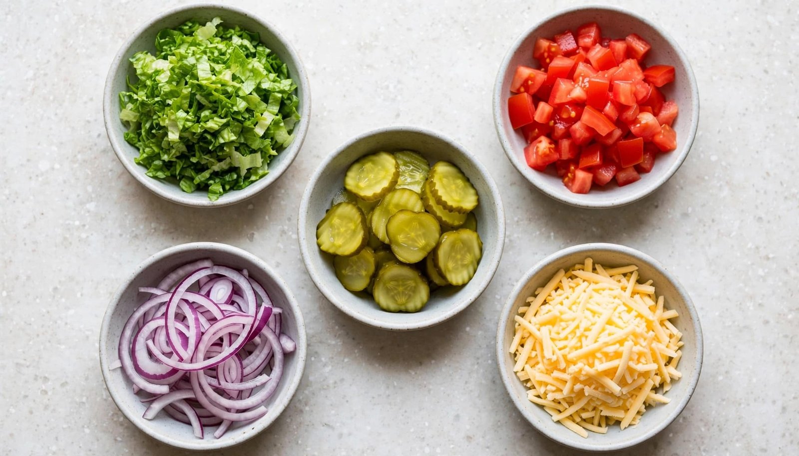 Overhead view of five small bowls containing chopped lettuce, diced tomatoes, pickles, sliced red onions, and shredded cheese.